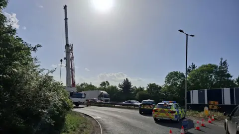 Norfolk Police A crane towering into the sky on the A11 in Thetford in Norfolk. Cones have been placed to stop traffic coming on to the road off a roundabout, and a fluorescent Norfolk Police car is also parked up to stop traffic getting through.