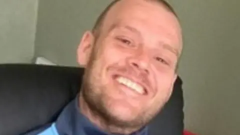 A man with very short hair in a blue tracksuit top sitting in a chair