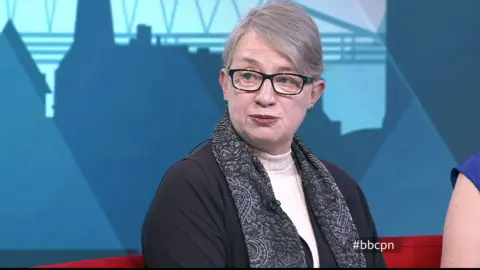 Natalie wears a white turtle neck sweater and dark blue cardigan with a paisley blue scarf. She is sitting on a red sofa in the studio.