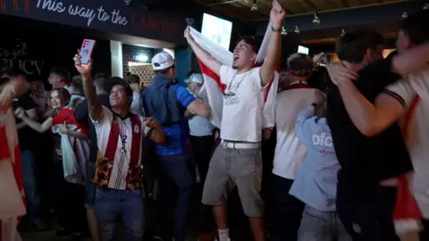 A large group of England football fans gathered in The Hyde Dendy pub in Paignton to watch the game against Mexico. They are celebrating a goal jumping up in the air. 