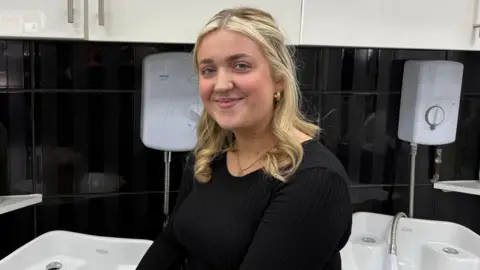 Ben Schofield/BBC Millie Clipston, standing by sinks in a hairdressers, wearing a black top, with long blonde hair. She has a nose piercing, and has in earrings and is wearing a necklace. 