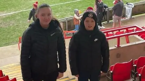 Siwan Davies Siwan and her brother standing in a football stadium with their back to the pitch and red seats on either side. They are both wearing black winter coats, Siwan with her hair tied back and her brother wearing a Wrexham bobble hat.