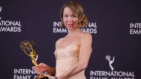 Getty Images Anna Maxwell-Martin stands in front of a backdrop covered in International Emmy branding. She is holding a large, shining gold trophy and is wearing a pale peach-coloured dress. She is looking directly at the camera, with a slight smile on her face.