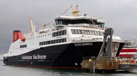 Glen Sannox beside a dock under a grey dawn sky. The boat has a line of flags along the top.