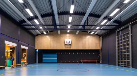 Getty Images The inside of a school gym, with climbing bars on the right hand side, packed away, and a basketball court and hoop taking up most of the hall.