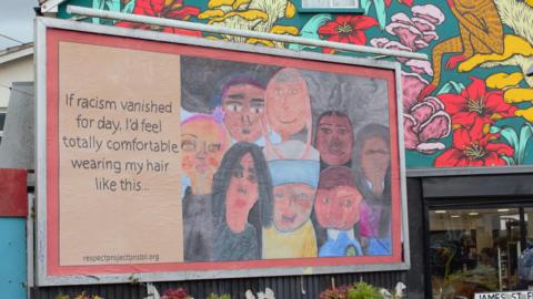 Image of the billboard. It has hand drawn portraits and text reading "if racism vanished for a day, I'd feel totally comfortable wearing my hair like this..."  