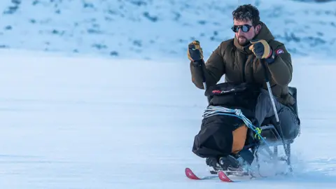 A man in an adapted sitting ski pulls himself using poles across snowy terrain. He is wearing a thick khaki jacket and sunglasses and is tied into the sit-ski with rope.