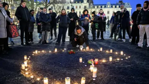 Uma dúzia de enlutados vestindo roupas de inverno reunidas em torno das flores e velas para homenagear as vítimas do tiroteio da Escola Ouribro. Ficou de pé, exceto por todas as pessoas trêmulas que decoram a vela