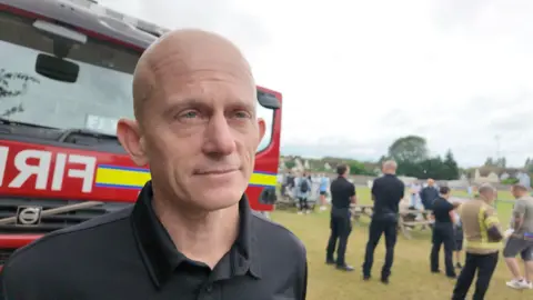 Oxfordshire's Chief Fire Officer, Rob MacDougall is wearing a black t-shirt and is standing in front of a red fire engine.