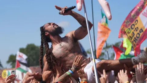 Bob Vylan crowd surfs during his performance at Glastonbury Festival. He is topless and is wearing white shorts and holding the mic in his left hand