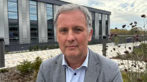 Ben Schofield/BBC Robert Evans is in the middle looking at the camera. He has short grey hair and is wearing a blue shirt and grey suit jacket. Behind him is a grey bricked building with tall windows.