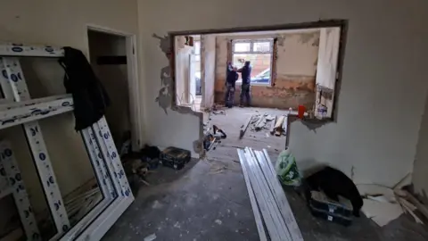 Bailey Capital Two workmen are fitting a new window frame into a hole in the far wall. The floorboards are exposed and other new window frames are resting against the wall to the left. The house looks in need of a lot of work.