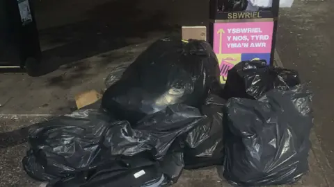 Darren Lloyd Several black bin bags dumped next to a public street bin that is full.