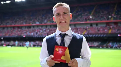 The FA / Getty Images A man is wearing a blazer, shirt and tie holding an award. He is stood on a football pitch, and behind him is a large stand in a stadium with many onlookers facing him. He is smiling and looking at the camera.