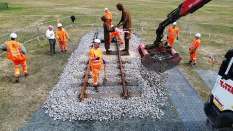 Daft As A Brush/Brian Burnie Construction workers wearing hi-vis clothing and hard hats work at the site of the "Forgotten Army" memorial in Marine Park, South Shields. The workers have laid a 10-metre length of railway track beneath a metal sculpture depicting Sunderland war veteran Len Gibson, handing a musical instrument to a child.