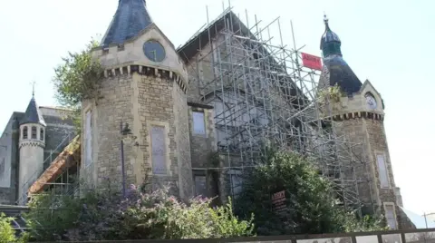 LDRS A Victorian building supported by scaffolding on the front. It has a pointed central roof with two turrets on each side of the main structure.