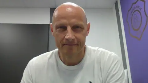 Matthias Heidrich A bald man smiles into the camera. He is wearing a white t-shirt. On his left, there is a purple wall with the badge of German football club Erzgebirge Aue.