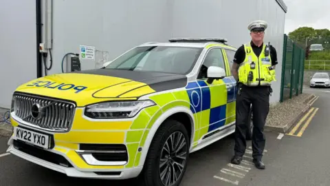DARREN WYlDE Stephen Hall a middle-aged man with large glasses wears a police uniform and stands next to a police car.