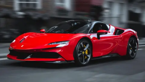 The Ferrari SF90 Stradale in bright red drives past brick buildings.