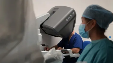 Shaun Whitmore/BBC Surgeon Nicholas Penney sits at a console for the da Vinci robot. His fingers are placed into controls mimicking his hands. The 3D visor encases his head, it is large and grey and has a da Vinci logo on the side. The shot looks through the arms of the robot and a member of theatre staff looks on to the right of the picture.
