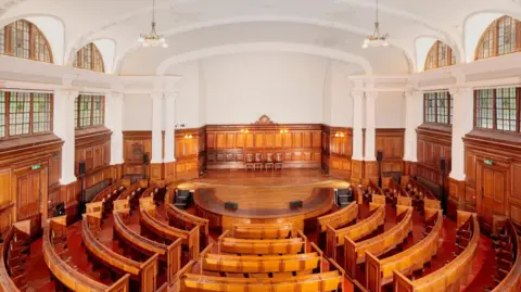 Historic England Archive Interior of a historic lecture hall featuring curved wooden benches facing a central stage, with dark wood panelling, tall arched windows, and an ornate white vaulted ceiling.