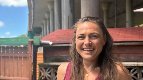 BBC A woman with long brown hair smiling wearing a red vest top standing in front of a wall with a motorway flyover in the background
