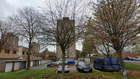 Google A car park with garages to the left and right. Vehicles are parked and there is a large concrete tower block in the background