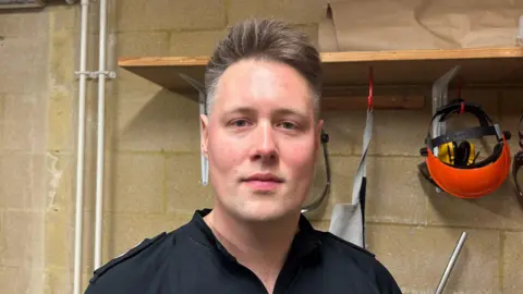 Ben Elliot looks at the camera in a windowless room with a brick wall and a row of helmets hung up under a shelf