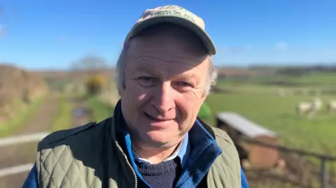 Michael is standing outside on his farm. He is smiling at the camera and is wearing a green cap. There are sheep in the field behind him. 