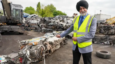 A man with a white beard and wearing a black turban, yellow hi-vis, grey suit jacket, blue shirt, blue tie and black trousers is standing in a scrapyard and pointing to a pile of crushed metal and debris. 