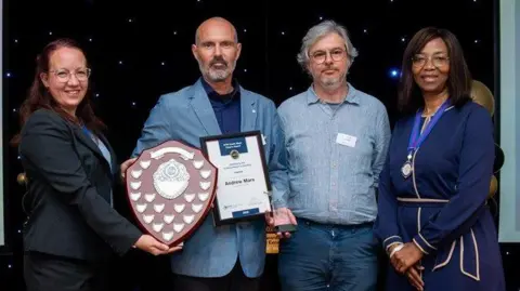 Andrew Marx (second left) receives the 'Unsung Planning Hero' award at Royal Town Planning Institute South West Awards for Planning Excellence. This includes a certificate and a large wooden shield. A man and two women are stood next to him.