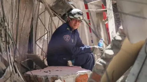 A rescue worker in a blue outfit and white helmet sits amid rubble