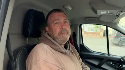 A man with short grey hair and beard , wearing a light brown hoodie, sitting in van.
