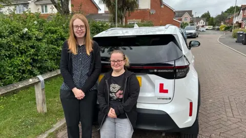 Chelsea Moon, who has a bone condition that stunts growth, waited a year for lessons with instructor Emma Hallett (left). Chelsea is just over four feet tall and is wearing a black hoodie, grey jeans and glasses. Emma, also in glasses, is wearing a black cardigan trousers and polka dotted shirt. They are standing behind a white car with learner plates.     