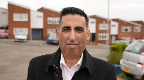 Qays Najm/BBC Ghulam Abbas, standing in a residential street, looking directly down the camera. He is smiling slightly and has short black hair. He is wearing a black overcoat, with an open-necked light-coloured shirt underneath. Behind him is a row of houses, with cars parked on several driveways. 