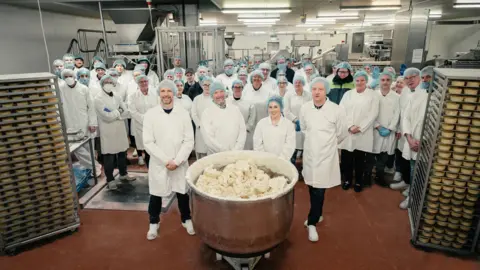 The floor of a bakery production centre, with dozens of employees standing for a group photograph alongside plenty of pastry