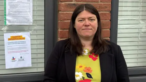 Emily Spurrell has long dark hair and is wearing a green floral patterned top and a black jacket. She is stood in front of the house window where a closure order has been attached to the window