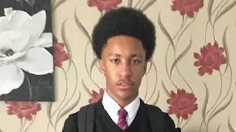 A picture of a teenage boy wearing a black school uniform, standing in front of floral wallpaper