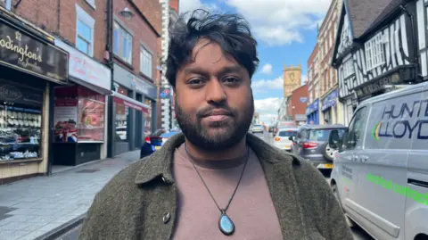 Sho Abdul is stood on Whitchurch high street and is looking into the camera. He is wearing a light brown T-shirt with an olive green wool jacket over the top, and is wearing a necklace with a large blue gemstone pendant. He has short dark hair and a short beard. Abdul is framed by shops along the edges of the photograph, with a church tower visible in the background. The sky is blue with some white clouds. 