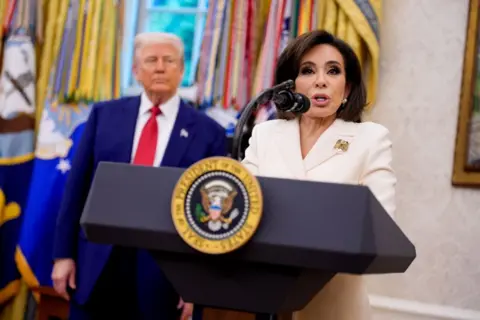Getty Images Jeanine Pirro está en el Despacho Oval hablando desde un podio con el escudo presidencial. Lleva un vestido blanco y un broche dorado en la solapa izquierda. Donald Trump está de pie detrás de ella, ligeramente desenfocado, con su característico traje azul marino y corbata roja.