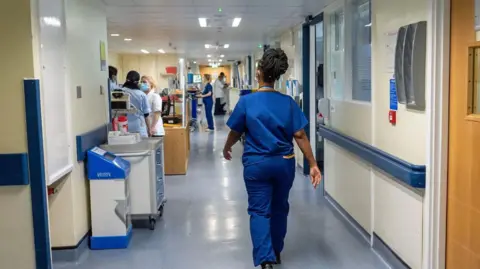 PA Media A nurse wears blue scrubs as she walks down a hospital corridor, there are other nurses in the background doing different tasks.