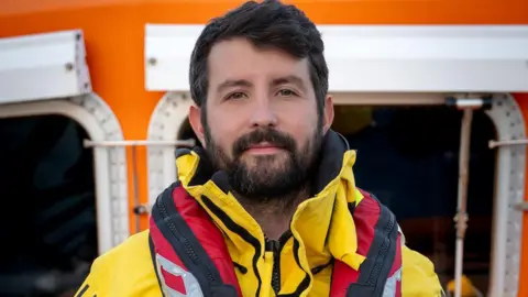 Will Roberts is smiling at the camera, wearing his RNLI lifeboat crew uniform which is a yellow anorak and a red lifebelt. He has black hair, a black beard and brown eyes. Behind him is the lifeboat wheelhouse which is orange with white trim.