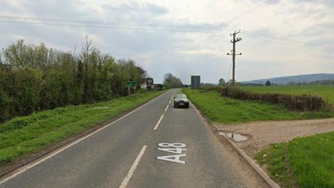 The A48 road with a black car driving towards the camera and a field on one side and trees on the other.
