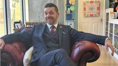 Ian Hyde is wearing a navy suit and is sitting on a red armchair in a room. He has short hair and a beard and is smiling.