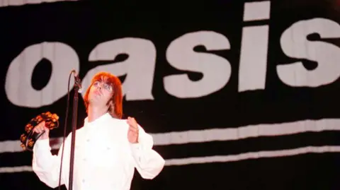 Getty Images Liam Gallagher holding a tambouring on stage in front of a giant Oasis banner at Knebworth in 1996