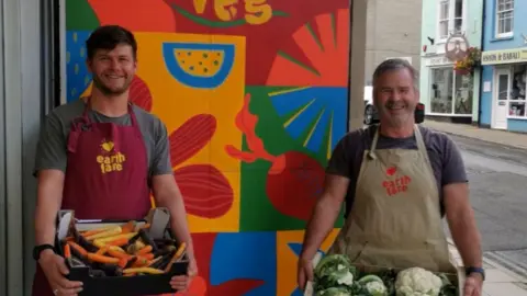 Henry Quinlan Henry and Andrew smile at the camera in front of a colourful wall. They are holding boxes of vegetables and wearing aprons with Earth Fare written on the front.