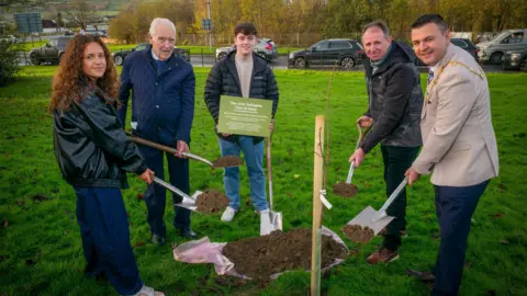 John's family members are pictured with mayor Ruairí McHugh holding shovels near the sapling. (L_R) Erin Gallagher, Jim Gallagher, Tiarnán Gallagher, John Donaghy and mayor Ruairí McHugh)
Mayor Ruairí McHugh) is wearing his mayoral chain in a cream suit jacket and navy trousers. Erin Gallagher (pictured far left) is in a dark leather jacket and navy trousers. She has long curly red hair. Jim Gallagher is in a navy coat and holding a plaque near the sapling. 