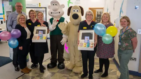 Wallace and Gromit characters wave at the camera on a ward at the Bristol Royal Children's Hospital. They are surrounded by members of staff, some of which are holding balloons and boxes of cupcakes.