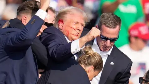 Donald Trump with blood on his right cheek and ear raises fist while surrounded by security staff