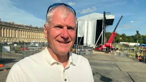 A man in a white polo shirt looks at the camera with a large stage behind and the Royal Crescent in Bath.